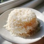 A perfectly molded mound of fluffy white thai coconut rice served on a simple white plate, illuminated by sunlight.