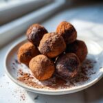 A pile of fudgy date brownie balls generously dusted with cocoa powder on a white plate.