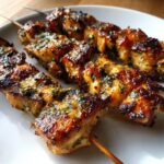 Three perfectly grilled garlic parmesan chicken skewers resting on a white plate, glistening with sauce and herbs.
