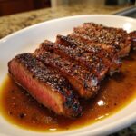 Slices of medium-rare garlic pepper steak resting in a rich brown sauce on a white plate.