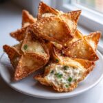 A stack of golden brown, crispy homemade crab rangoons piled on a small white plate, one showing the creamy filling.