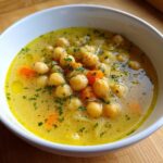 Close-up of a white bowl filled with bright yellow Greek lemon chickpea soup, featuring whole chickpeas, diced carrots, and fresh parsley.
