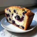 A thick slice of moist blueberry bread featuring a prominent cream cheese swirl and baked blueberries, served on a white plate.