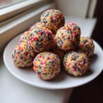 A pile of funfetti-style no bake cake batter protein balls coated in rainbow sprinkles on a white plate.