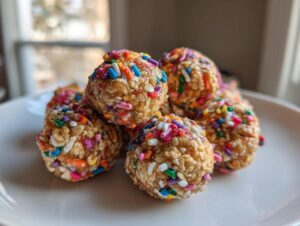 A stack of no bake cake batter protein balls coated in colorful rainbow sprinkles on a white plate.