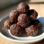 A stack of rich, dark no bake hazelnut cacao bites speckled with lighter nuts and oats, sitting on a small white plate.