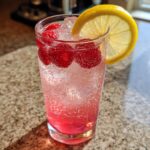 Close-up of a tall glass containing a bubbly pink lemonade mocktail garnished with fresh raspberries and a lemon slice.
