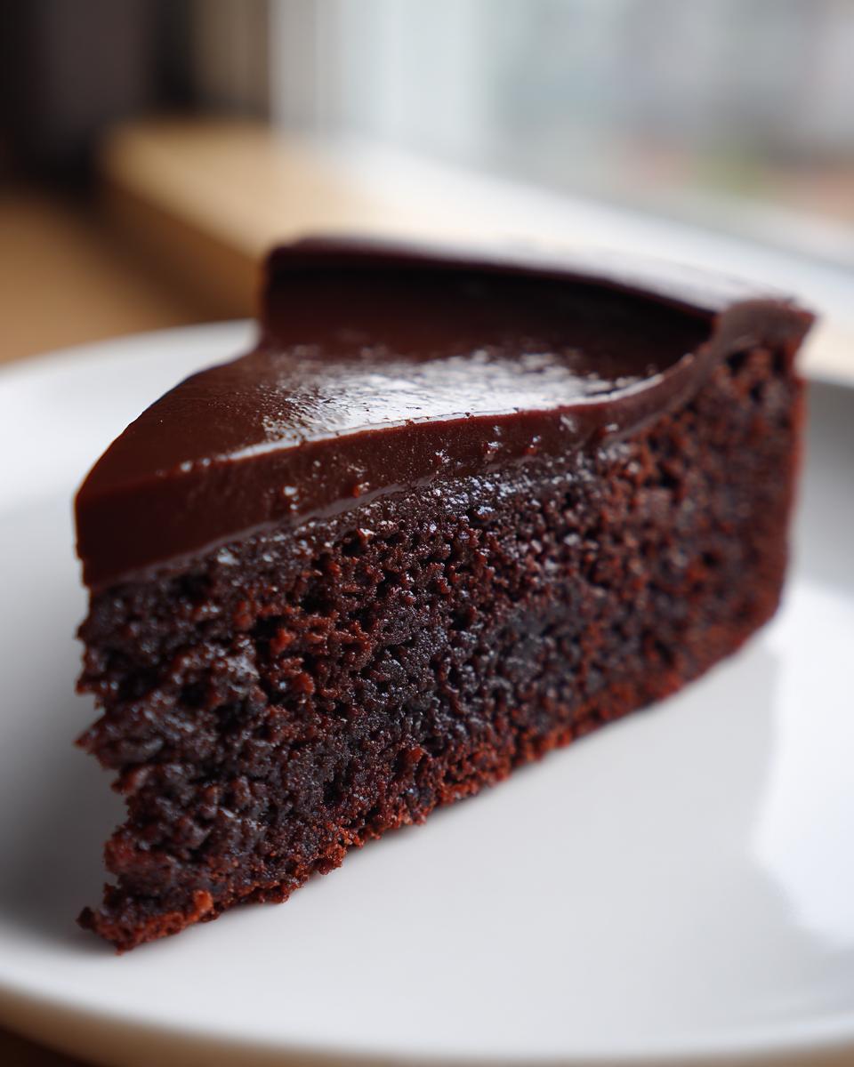 A decadent slice of flourless chocolate cake with a glossy chocolate ganache topping, served on a white plate.