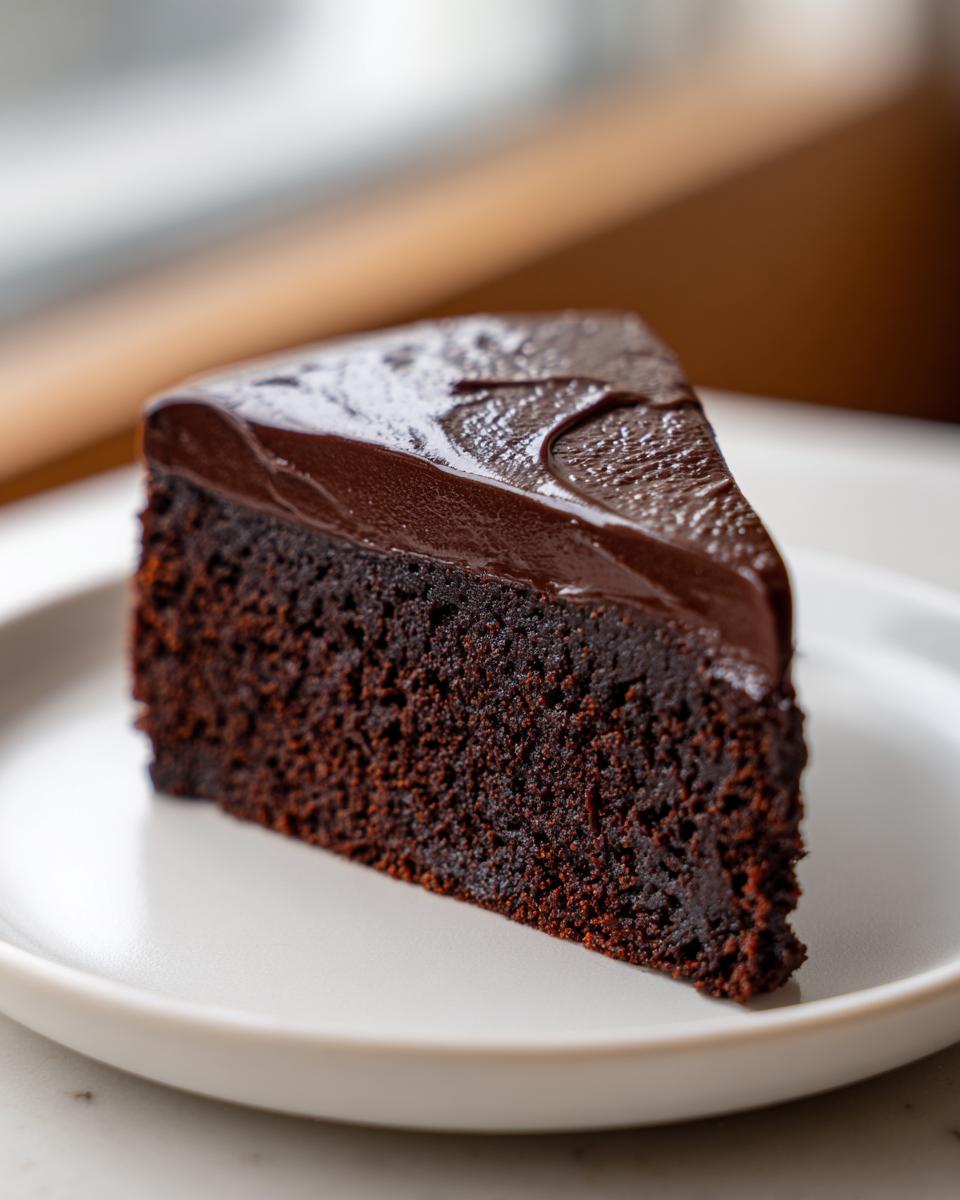 A decadent slice of flourless chocolate cake topped with shiny chocolate ganache, served on a white plate.