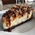 A decadent slice of Samoa Cheesecake featuring a dark crust, creamy filling, caramel, toasted coconut, and chocolate drizzle.