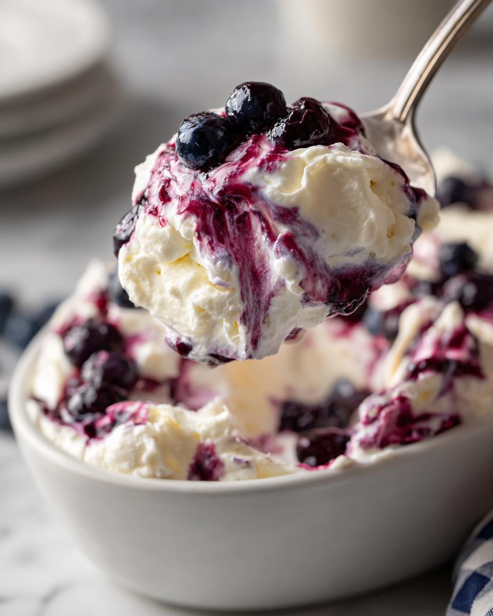A spoonful of creamy blueberry fluff salad topped with fresh blueberries and sauce being lifted from a white bowl.