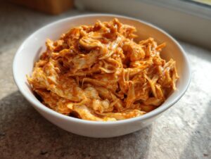 A white bowl filled with shredded chicken coated in a creamy, orange buffalo sauce, ready for slow cooker buffalo chicken chili.