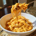 A spoonful of creamy, shredded buffalo chicken being lifted from a white bowl of slow cooker buffalo chicken chili.