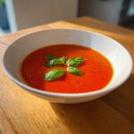 Close-up of a vibrant red bowl of simple tomato basil soup topped with fresh basil leaves.
