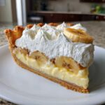 A perfect slice of banana cream pie recipe showing layers of crust, custard, bananas, and whipped cream topping.