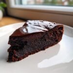 A close-up slice of dense, dark flourless chocolate cake topped with glossy chocolate ganache.