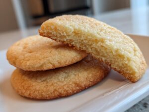 A stack of golden-brown sourdough sugar cookies, with one cookie broken in half showing the soft, cakey interior.