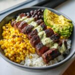 Close-up of a steak avocado and roasted corn bowl featuring sliced steak over rice, topped with creamy sauce.