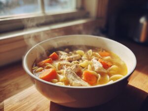 A steaming white bowl filled with rich broth, shredded chicken, egg noodles, and bright orange carrots, representing slow cooker chicken noodle soup.