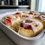 Close-up of gooey, freshly baked strawberry cinnamon rolls topped with white icing in a white baking dish.