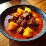 Close-up of a bowl of hearty goulash recipe featuring tender beef chunks and large pieces of yellow potato in a rich, red sauce.