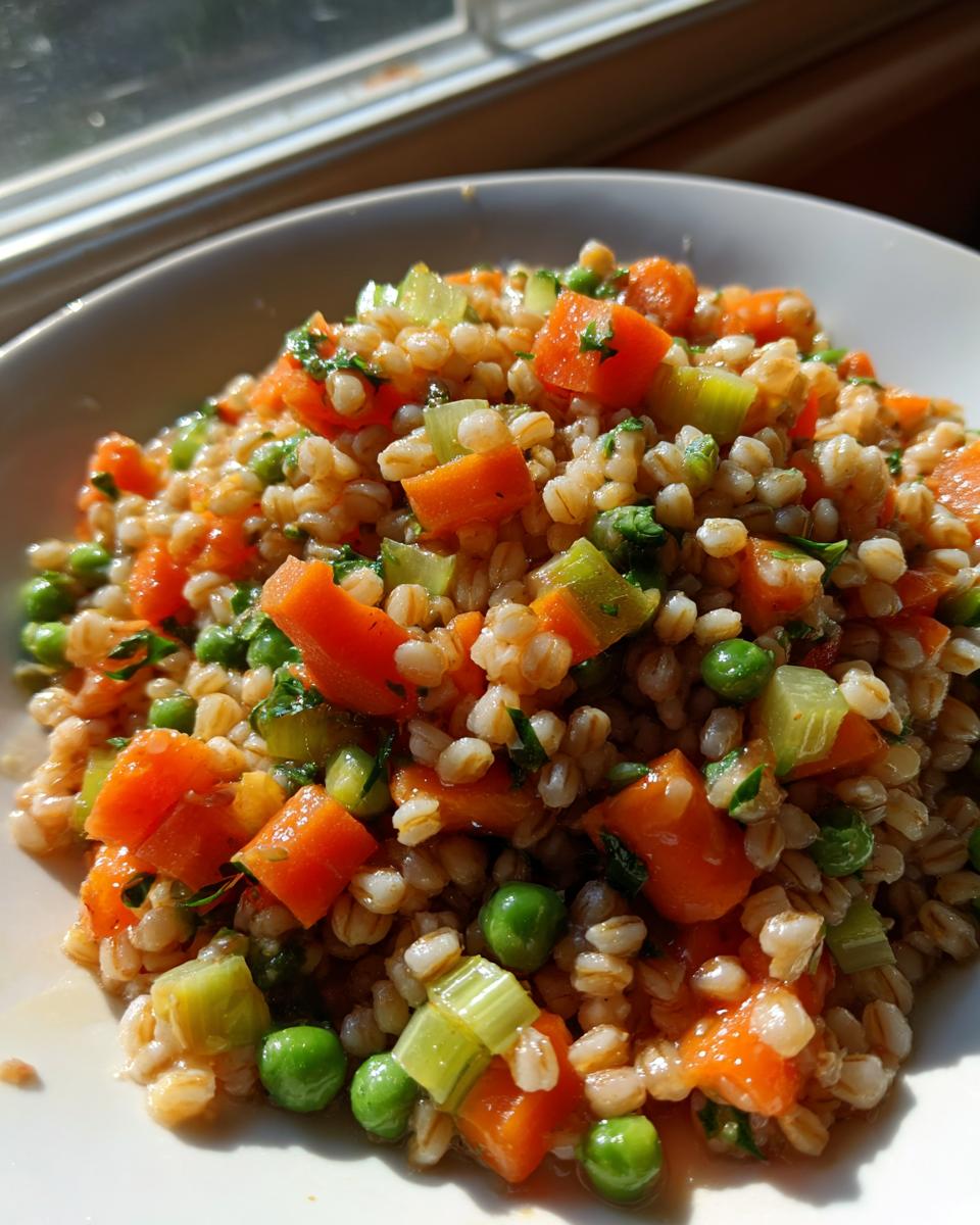 A close-up shot of a hearty vegan vegetable barley salad featuring plump barley grains, bright orange carrots, green peas, and celery.