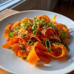 Close-up of a vibrant raw carrot salad made with shaved carrot ribbons, topped with sesame seeds and sliced green onions.