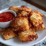 Close-up of perfectly crispy, golden-brown air fryer toasted ravioli piled on a white plate next to a small bowl of marinara sauce.