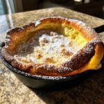 A golden, puffed Dutch baby pancake dusted heavily with powdered sugar, served in a black cast iron skillet.