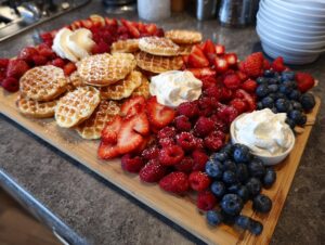 A vibrant Breakfast grazing board featuring mini waffles dusted with powdered sugar, fresh strawberries, raspberries, blueberries, and whipped cream.