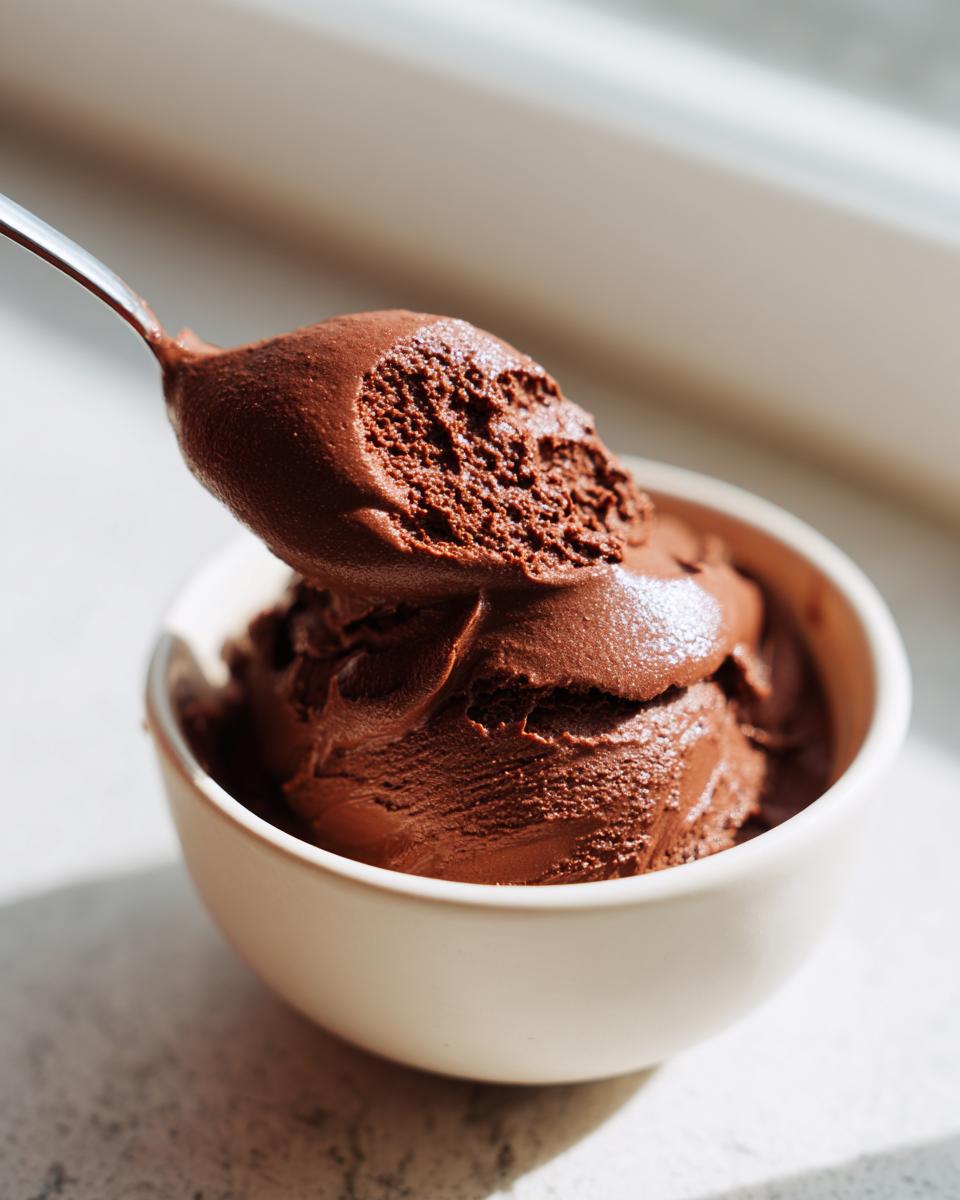 A spoonful of rich, dark chocolate cottage cheese mousse being lifted from a small white bowl.