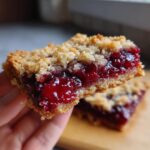 A hand holding a square bar of classic cherry cobbler showing the thick, bright red cherry filling and crumb topping.