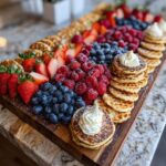 A stunning, colorful breakfast grazing board featuring stacks of mini pancakes with whipped cream, strawberries, blueberries, and raspberries.