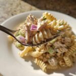 A spoonful of creamy tuna pasta salad featuring fusilli pasta, tuna chunks, and red onion, lifted above a white plate.