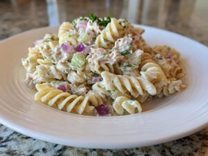 A close-up of a serving of creamy tuna pasta salad made with rotini pasta, red onion, and celery on a white plate.