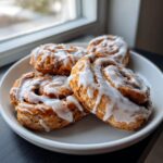 Four freshly baked Protein cinnamon rolls generously drizzled with white icing, served on a white plate near a window.
