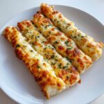 Four golden brown, cheesy garlic cheesy breadsticks topped with parsley served on a white plate.