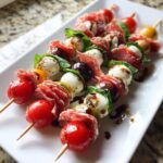 Close-up of several gourmet antipasto skewers featuring cherry tomatoes, mozzarella balls, salami, basil, and olives drizzled with balsamic glaze.