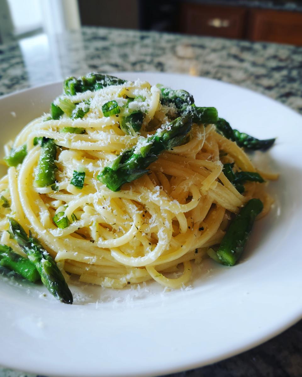 A close-up of a white plate filled with Lemon Asparagus Pasta, topped with grated Parmesan cheese and fresh green asparagus pieces.
