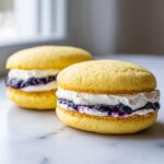 Two delicious Lemon blueberry whoopie pies featuring bright yellow cakes, white cream, and dark blueberry filling on a marble surface.