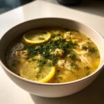 Close-up of a bowl of bright Mediterranean lemon chicken soup garnished with fresh dill and lemon slices.