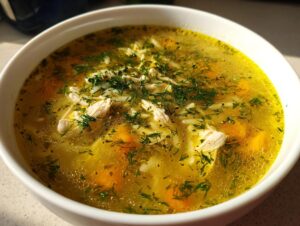 Close-up of a white bowl filled with bright yellow Mediterranean lemon chicken soup, featuring shredded chicken, carrots, rice, and fresh dill.