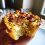 Close-up of a hand holding a Mini quiche lorraine with a bite taken out, showing the fluffy interior and bacon topping.
