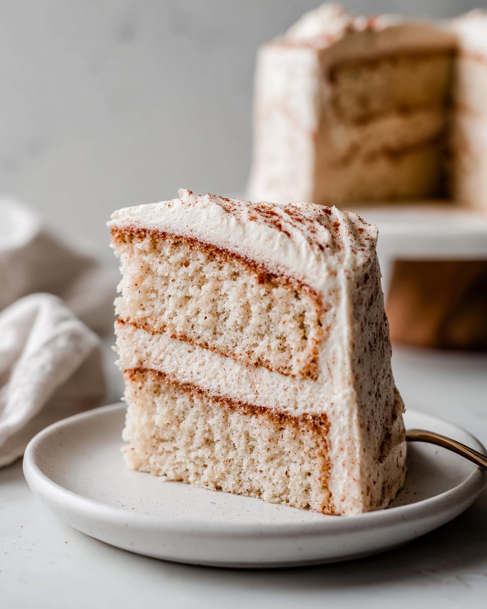 A tall slice of moist Snickerdoodle cake with cinnamon swirl layers and cream cheese frosting on a white plate.