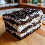 A rich, layered slice of Oreo icebox cake featuring dark chocolate cookie layers and white cream filling, topped with crumbs.