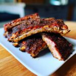 Close-up of tender Oven baked ribs heavily coated in a dark, flavorful dry rub, stacked on a white plate.