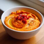 Close-up of creamy, bright orange roasted red pepper hummus topped with olive oil and chopped sun-dried tomatoes.