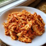 Close-up of a mound of juicy, shredded salsa chicken coated in red sauce served on a white plate.