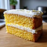 A thick slice of bright yellow Lemon cake with cream cheese frosting layered between the two sponge layers and on top.