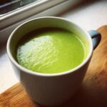 A close-up of vibrant green Spring pea soup served hot in a white mug, resting on a wooden surface.